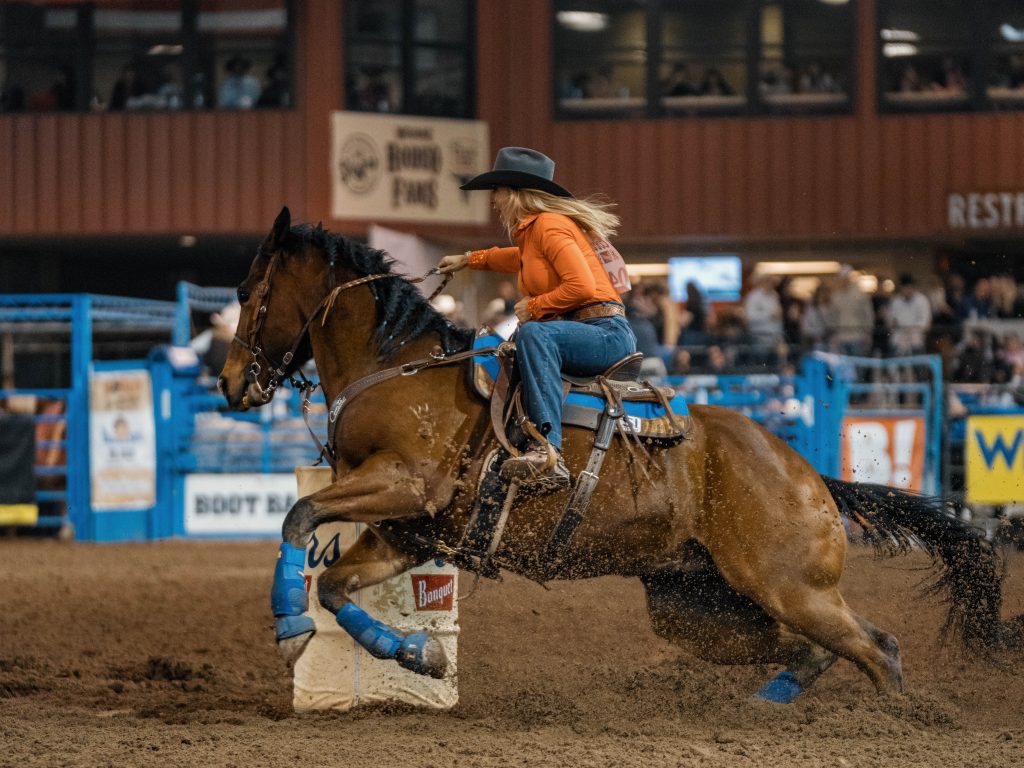 A female barrel racer wearing an orange long-sleeve shirt, jeans, and a black cowboy hat leans into a tight turn around a white barrel during an indoor rodeo competition. Her bay horse kicks up a spray of dirt as it hugs the barrel, blue sport boots on its legs. A crowded arena and sponsor banners are visible in the background.